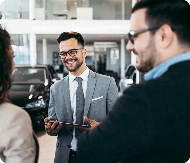 Un vendeur souriant en costume et lunettes tenant une tablette, parlant à deux clients dans un concessionnaire automobile