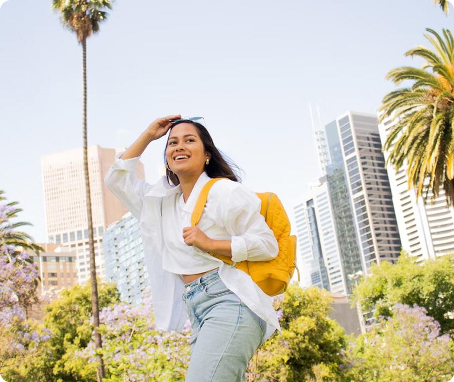 Une jeune femme souriante avec un sac à dos jaune, le bras levé pour protéger ses yeux du soleil, dans un parc urbain