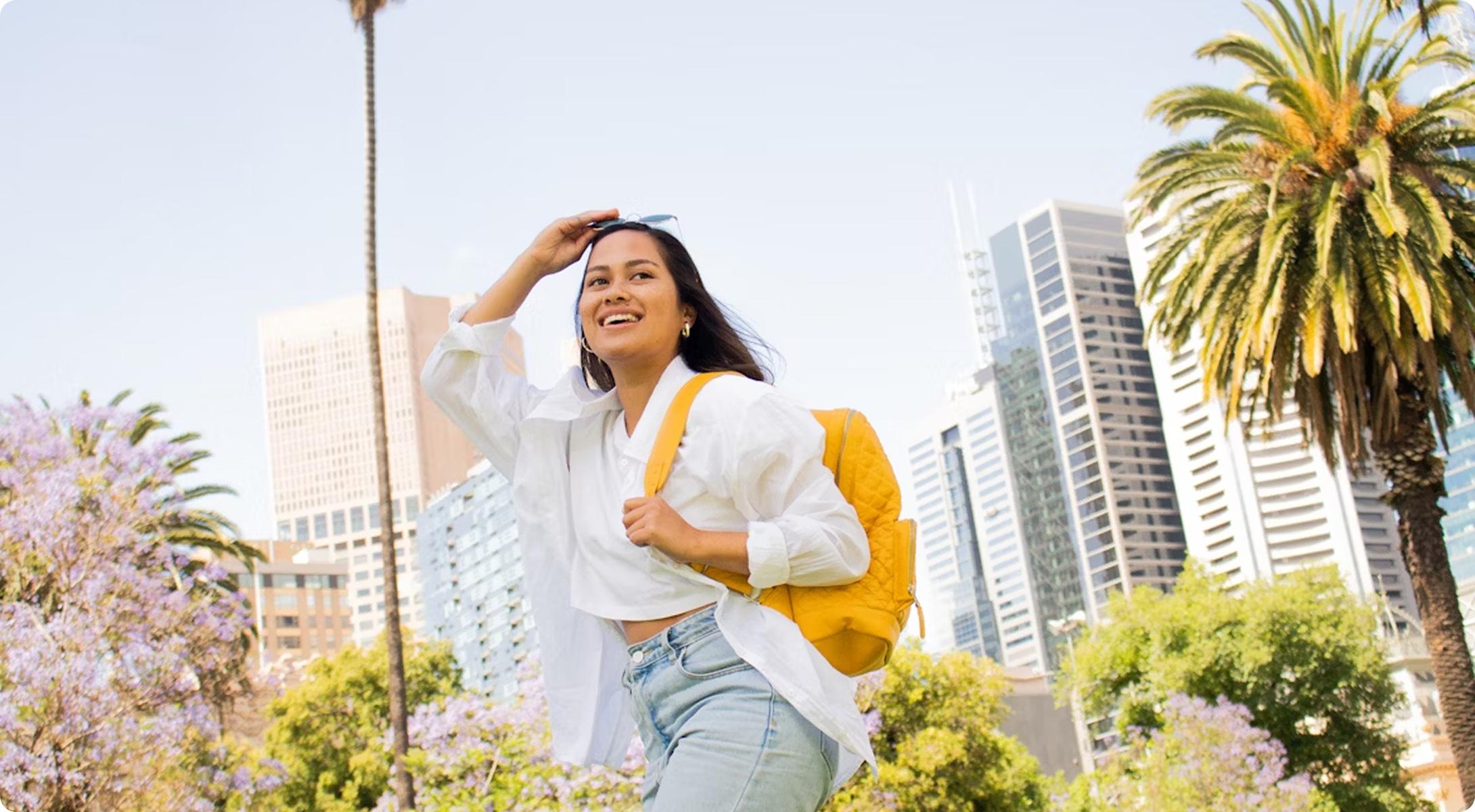 Une jeune femme souriante avec un sac à dos jaune, le bras levé pour protéger ses yeux du soleil, dans un parc urbain