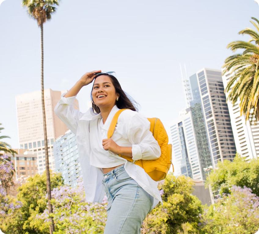Une jeune femme souriante avec un sac à dos jaune, le bras levé pour protéger ses yeux du soleil, dans un parc urbain